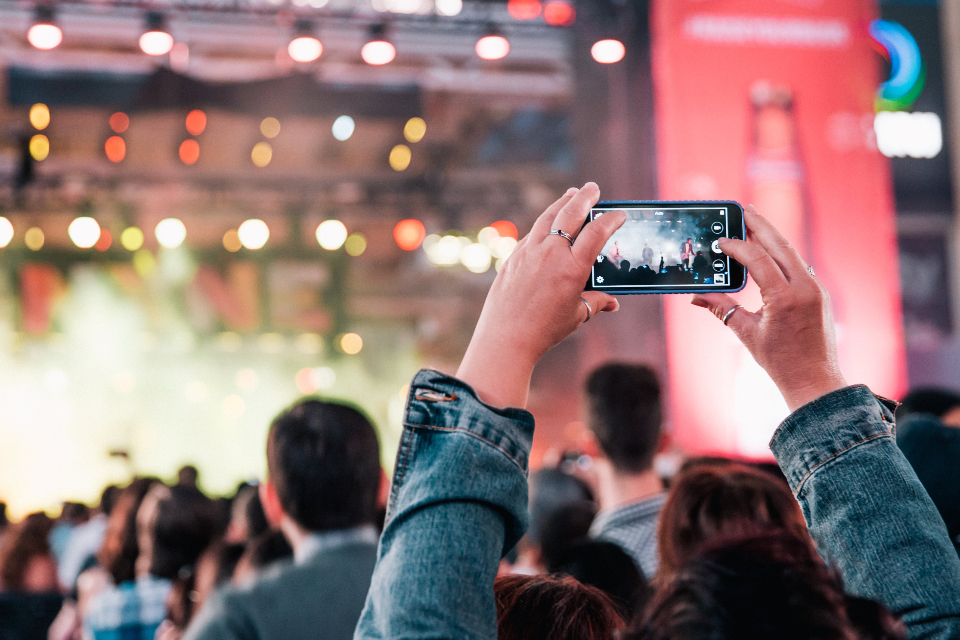 A concert crowd seen from behind, one person holding a phone up to record the stage.